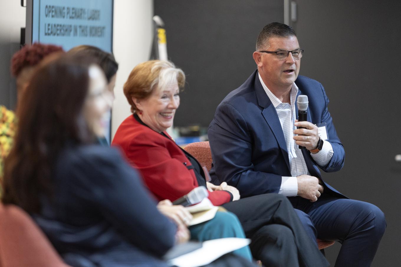 Man and woman on stage smiling while speaking to unseen group ahead