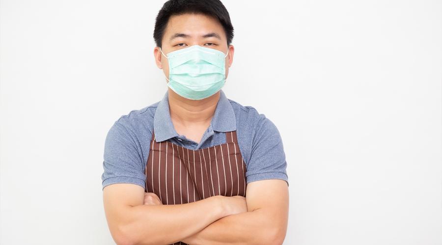 Asian male wearing an apron and a medical face mask, standing with his arms crossed.