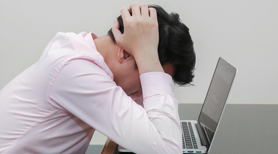 A manin a white dress shirt leans over his laptop computer, elbows on a table, with hands clenched behinds his head.