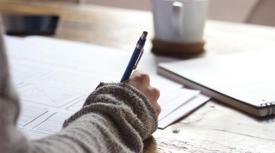 person writing on brown wooden table near white ceramic mug