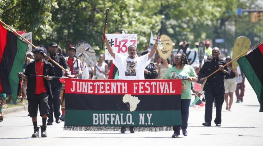 A group marches behind a sign that reads "Juneteenth Festival Buffalo, NY with an image of the African continent. Marchers hold up Black Liberation Flags and large cutout drawings of African masks. 