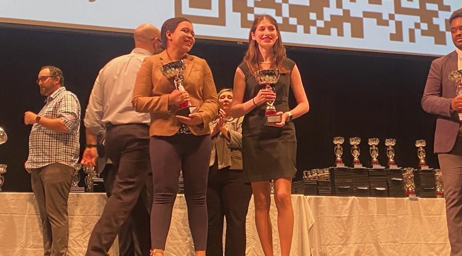 Two students stand on a stage holding trophies