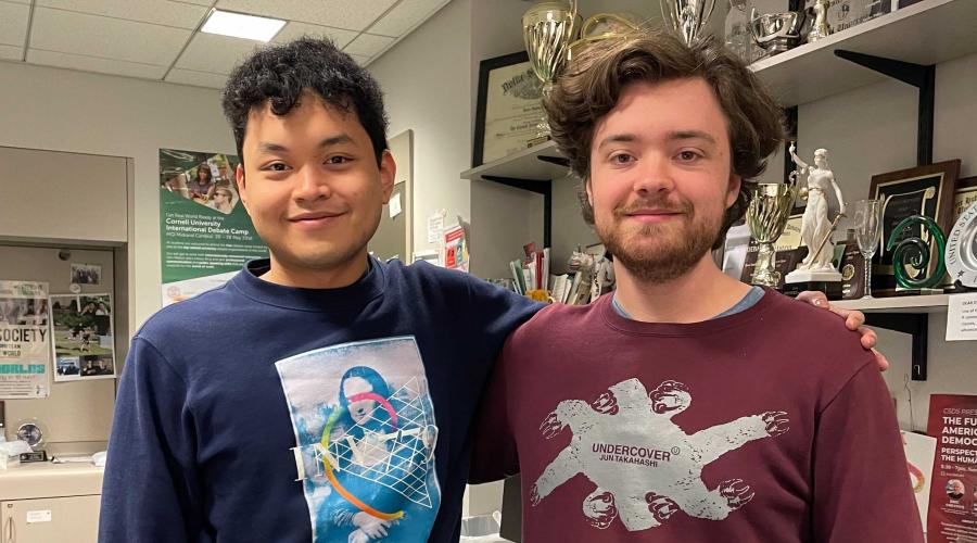 Two students stand side by side with an arm around each other in front of a wall of trophies