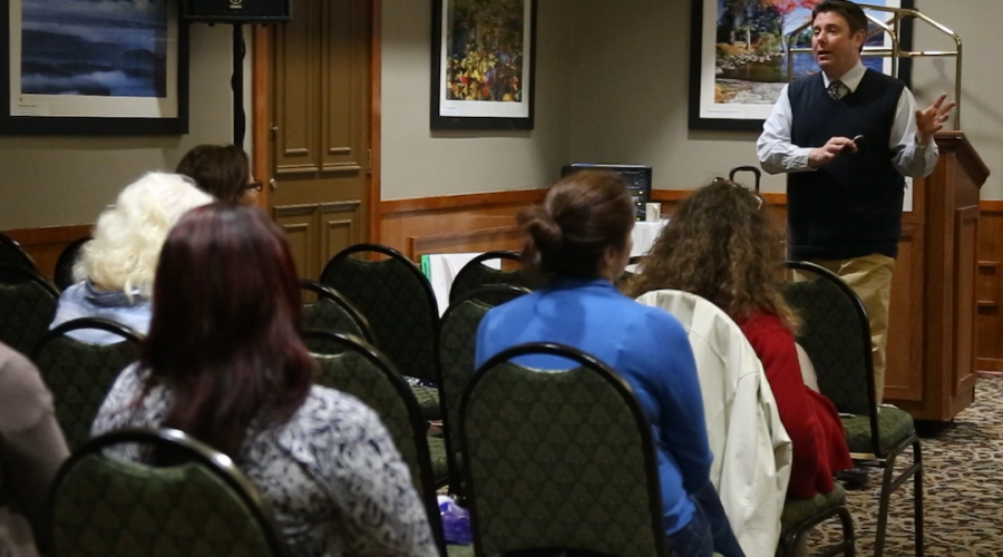 Instructor Jeffrey Tamburo wears a long sleeve light blue button up shirt with a necktie and blue sweater vest and stands in front of a podium while lecturing seated participants 