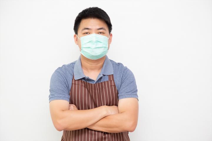 Asian male wearing an apron and a medical face mask, standing with his arms crossed.