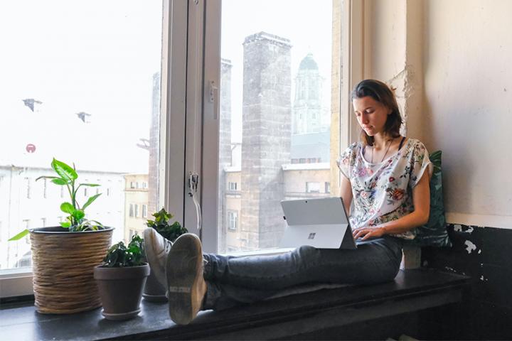 Young woman working on a laptop, sitting on a window sill.