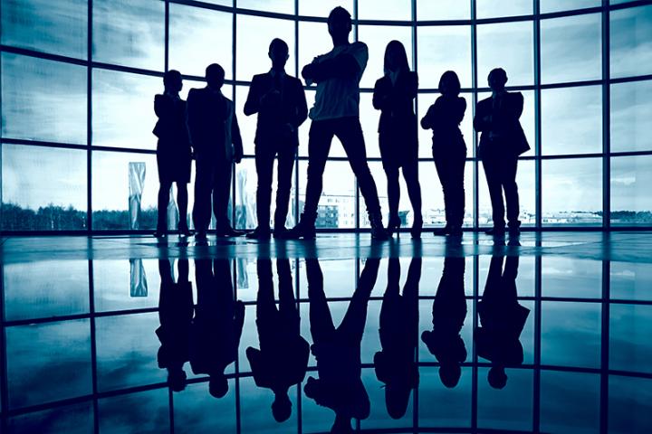 A group of seven people stand in front of floor to ceiling windows with blue skies beyond.