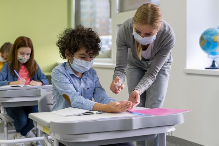 A female teacher wearing a mask puts hand sanitizer on a student