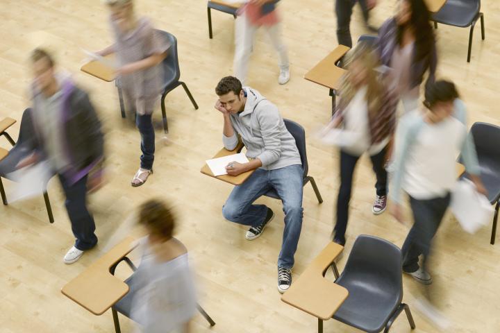 A college student sits at a desk while his classmates rush around