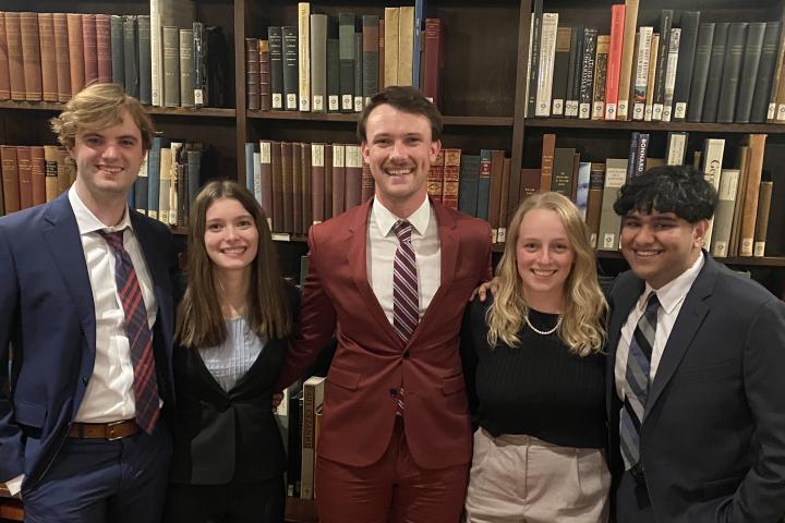 5 Cornell representatives in dress clothes stand in front of a book case