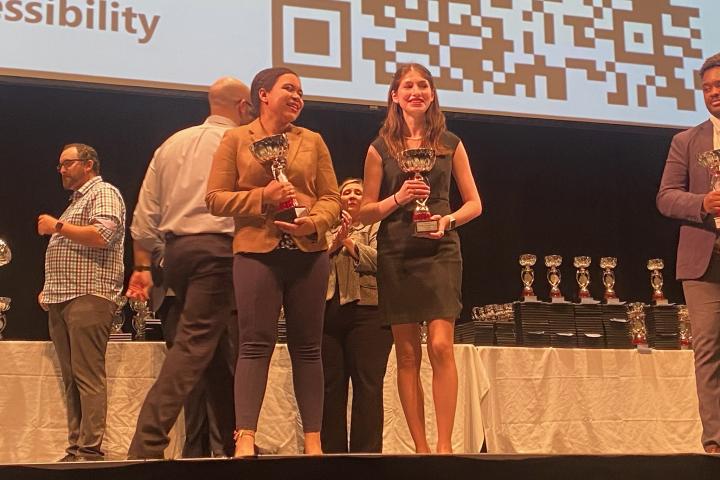 Two students stand on a stage holding trophies
