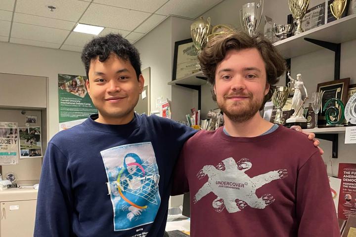 Two students stand side by side with an arm around each other in front of a wall of trophies