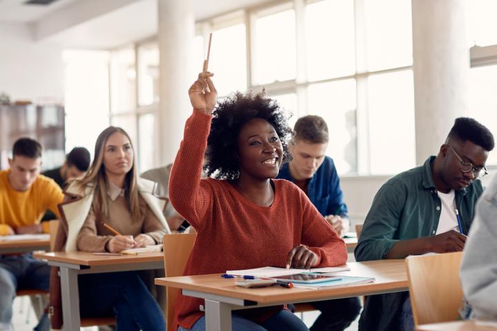 A high school classroom with students sitting at their desks while listening to a lecture. A student in the center of the image has raised a hand and is smiling while waiting to be called on.