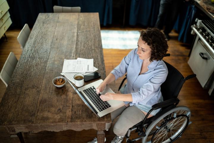 Photo of woman in a wheelchair at a table with laptop and letters beside her.
