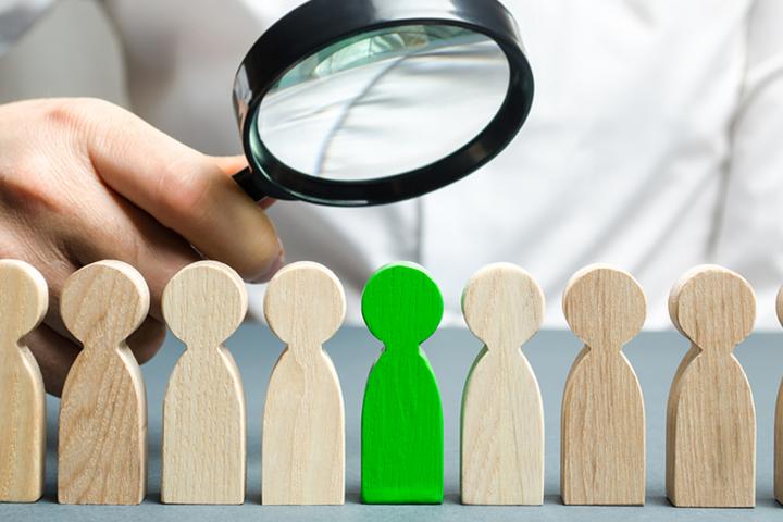 Wood figures in a row with a single green one being singled out by a businessman with a magnifying glass.