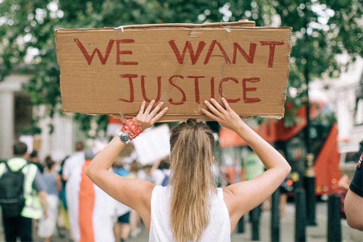 A young woman at a protest holds a sign that reads "We Want Justice"