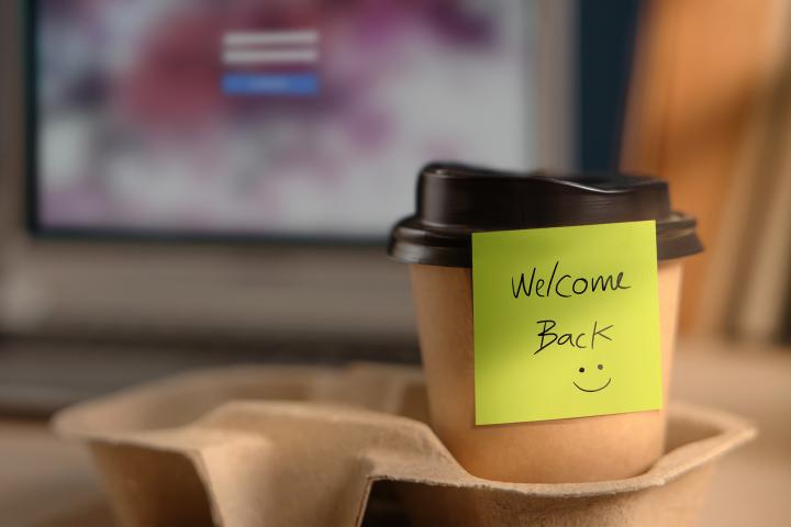 A post-it note that says "Welcome Back" sticks to a take out coffee cup in front of a computer.